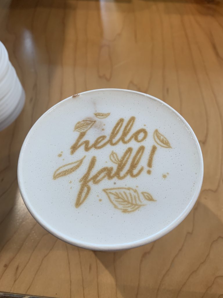 Latte with printed message "hello fall!" surrounded by leaf designs on a wood counter.
