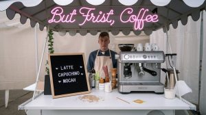 A fake coffee cart setup with a Breville machine, crooked letterboard menu with misspelled drink names, and a neon sign that says “But Frist, Coffee.”